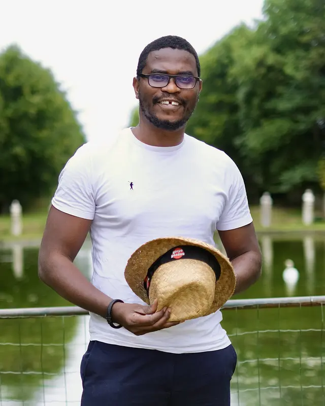 Smiling lecturer outdoors in glasses and a white T-shirt, holding a straw hat, with a pond and trees behind